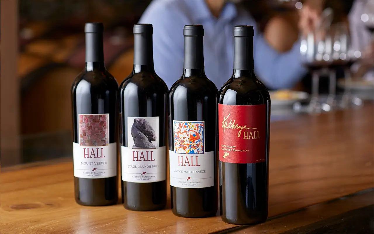 Four bottles of HALL Napa Valley Cabernet Sauvignon, including Mount Veeder, Stags Leap District, Jack's Masterpiece, and Kathryn Hall, displayed on a wooden bar for the Primary Colors wine club.