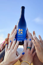 Bottle of Baca Zinfandel held aloft by multiple people with a clear blue sky background