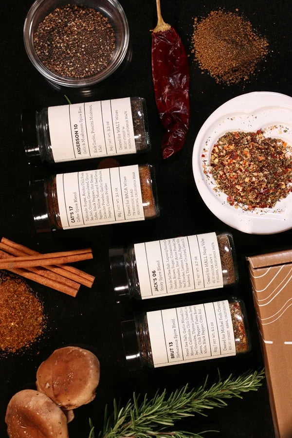 Image showing the 4 spice jars included int the HALL Family Spice Boz Set. The Jars are flatlayed on a black countertop surrounded by the box, a pinch bowl of spice, cinnamon sticks, dough a dried pepper and rosemary.