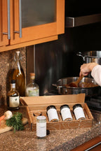 The HALL Family Spice Box displayed on a granite countertop next to a woman cooking.