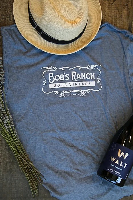 Flat Lay image of a grey-blue WALT Wines t-shirt featuring the Bob's Ranch Vineyard name and 2023 Vintage. The image also contains a bottle of WALT 2023 Bob's Ranch Pinot Noir, a woven hat and bunch of dried lavender.