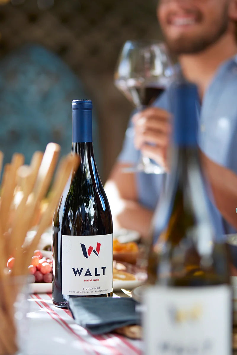 Bottle of Walt Sierra Mar against a blurred background of a man drinking a glass of red wine.