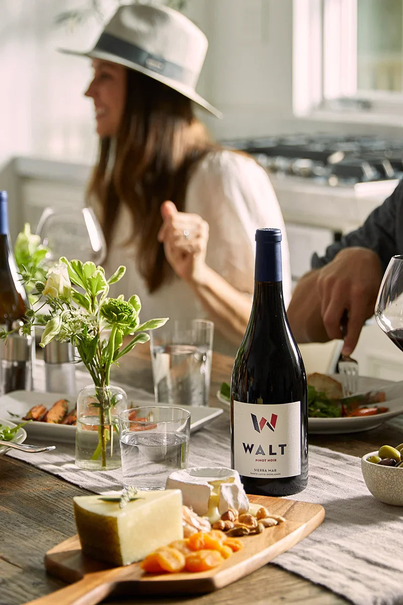Bottle of 2024 WALT Sierra Mar Pinot Noir on a table with a cheese plate and people eating food.